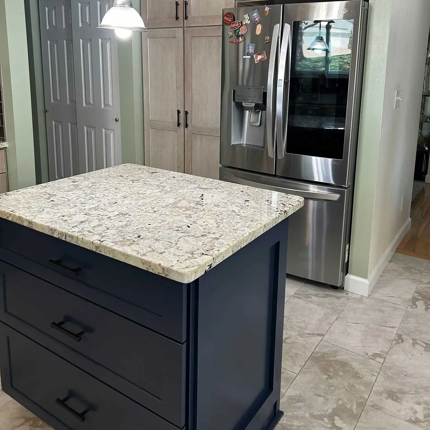 A navy blue kitchen island with a speckled granite countertop in front of wooden cabinets and a stainless refrigerator.