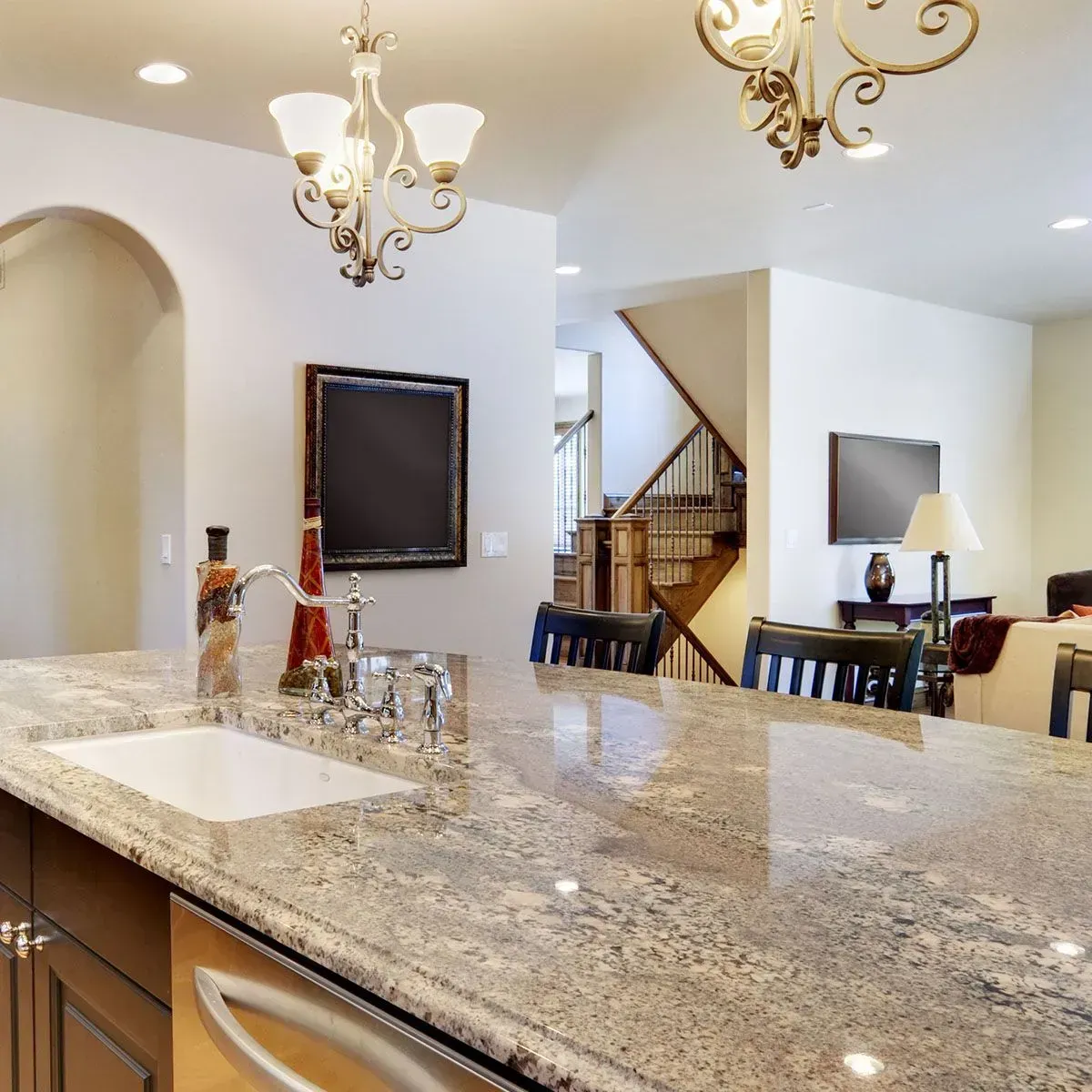A kitchen island with a granite countertop and stainless steel faucet, with a staircase and living area in the background.