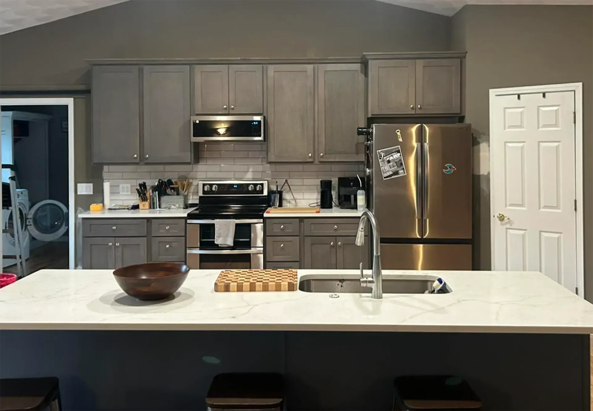 A modern kitchen with grey cabinets, stainless steel appliances, and a large white island with a sink and cutting board.