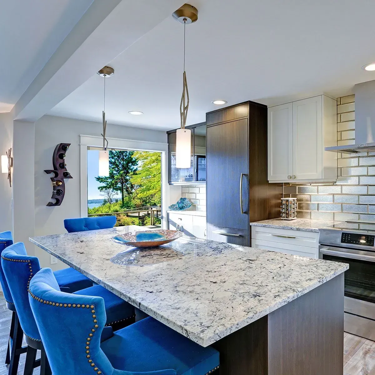 A bright, modern kitchen featuring a granite-topped island with blue upholstered chairs and a view of trees and water.
