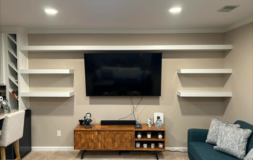 A wall-mounted TV above a wooden media console, flanked by white floating shelves against a beige wall in a living room.