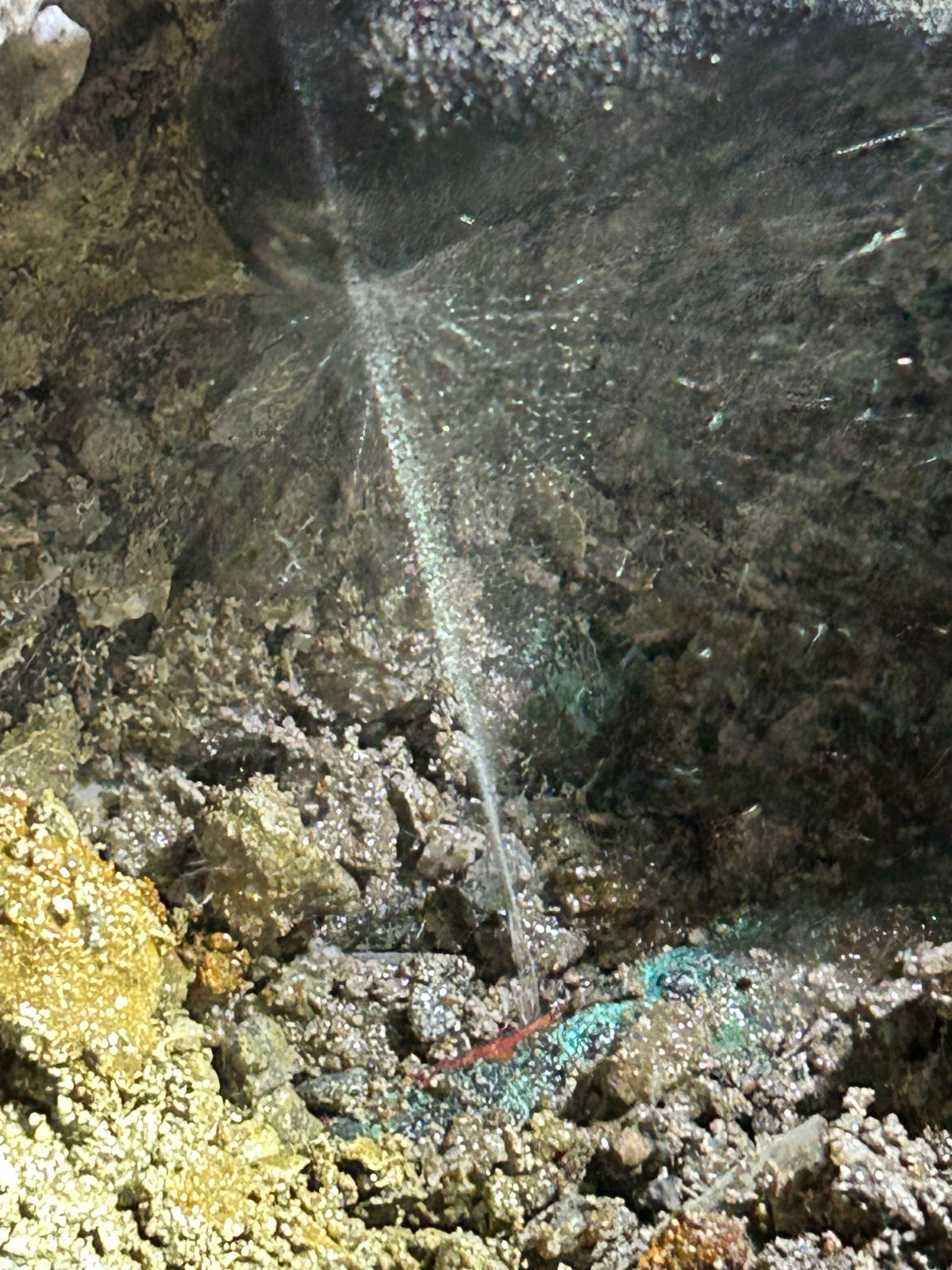 A pressurized water leak spraying from a pipe buried in dirt and rocky soil.