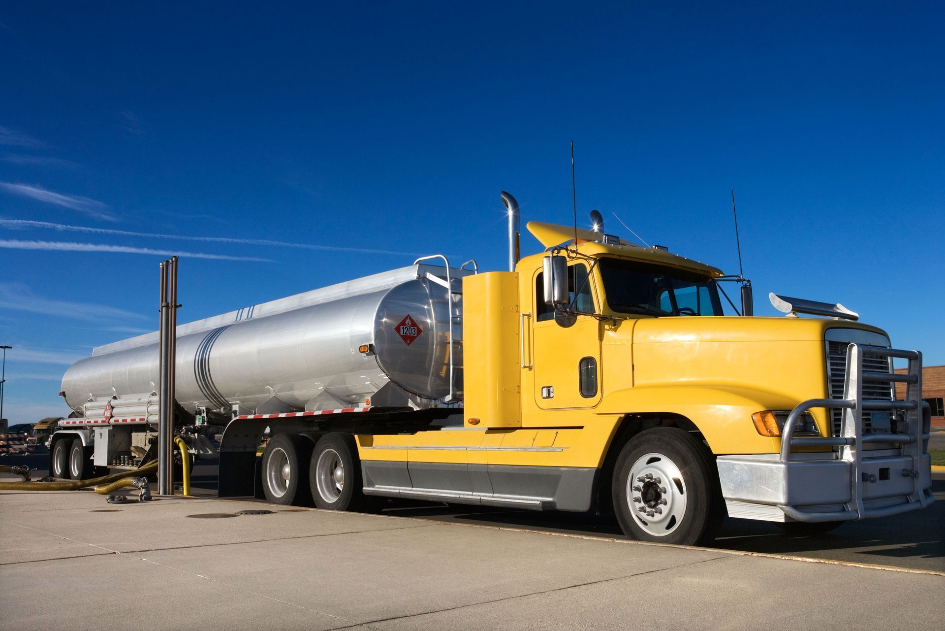 A yellow semi-truck with a silver tanker trailer parked on an asphalt lot under a clear blue sky.