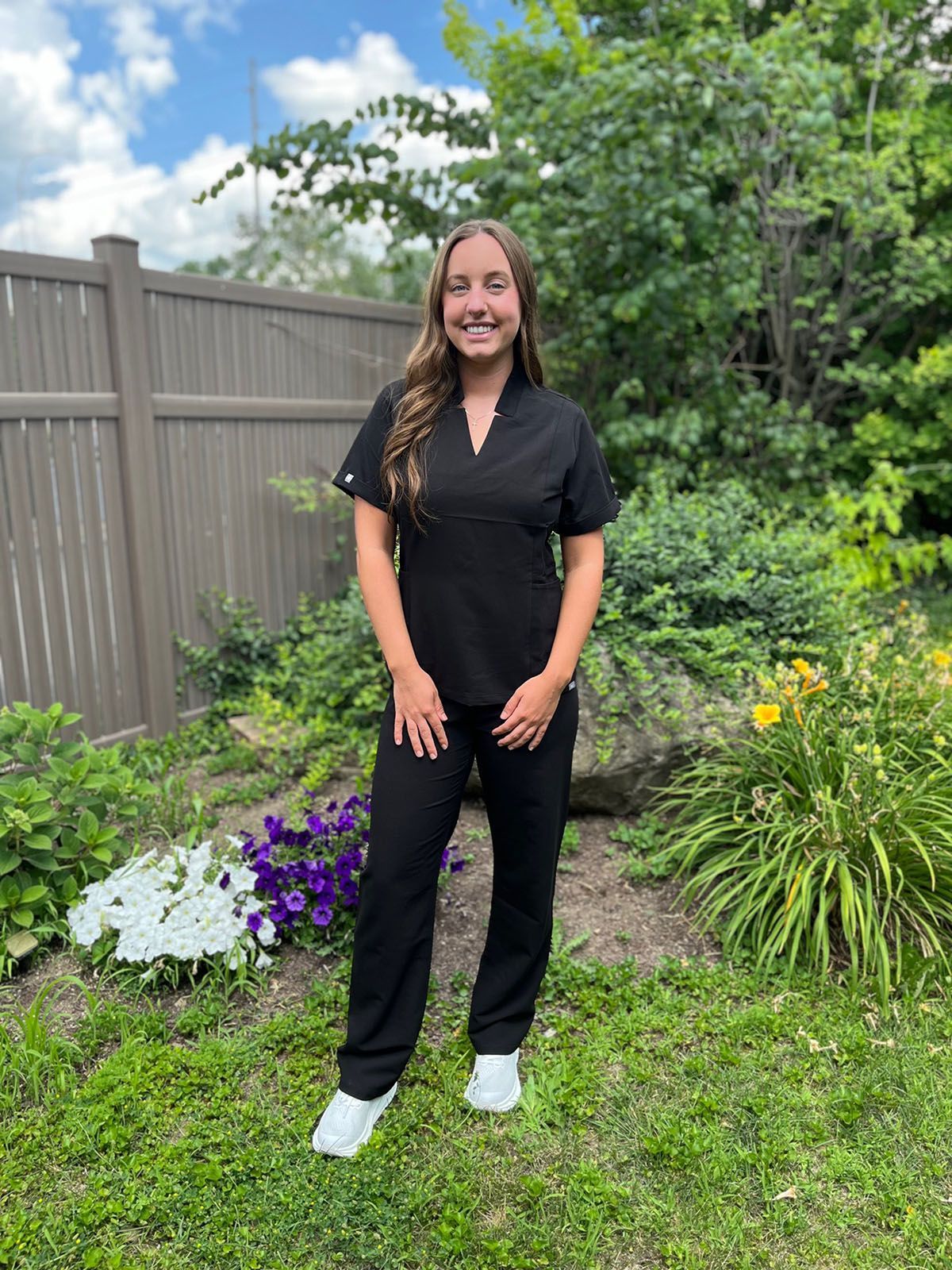 Woman in black scrubs and jacket stands outdoors in a garden, smiling.