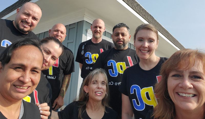 Group of people wearing matching black shirts with a colorful logo, smiling in front of a building.