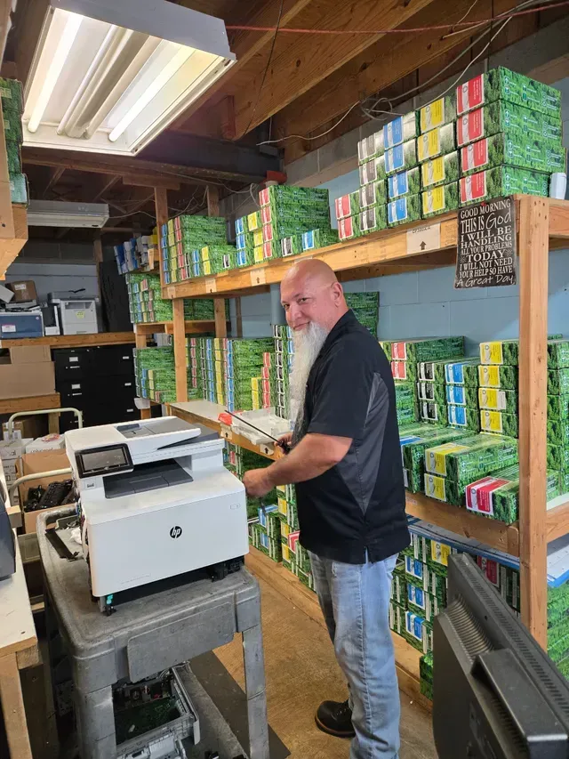 Man with a white beard holds something by a printer. Shelves of boxes are behind him in a cluttered storage space.