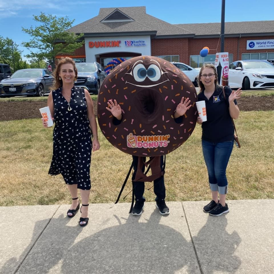 Two women and a Dunkin' Donuts mascot pose in front of a Dunkin' Donuts store. The mascot is a chocolate donut with arms and legs.