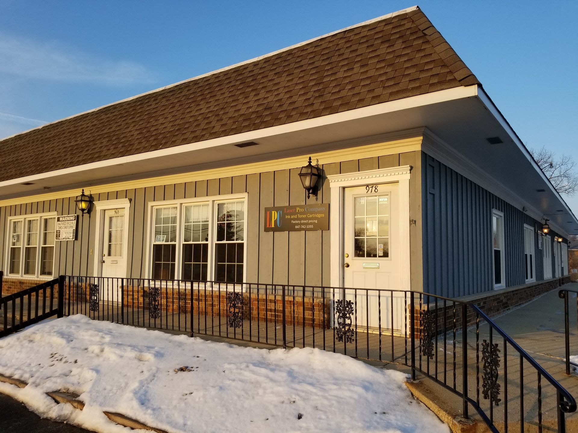 Exterior of a gray building with a brown roof and snow on the ground; a brick base and decorative wrought iron railing.