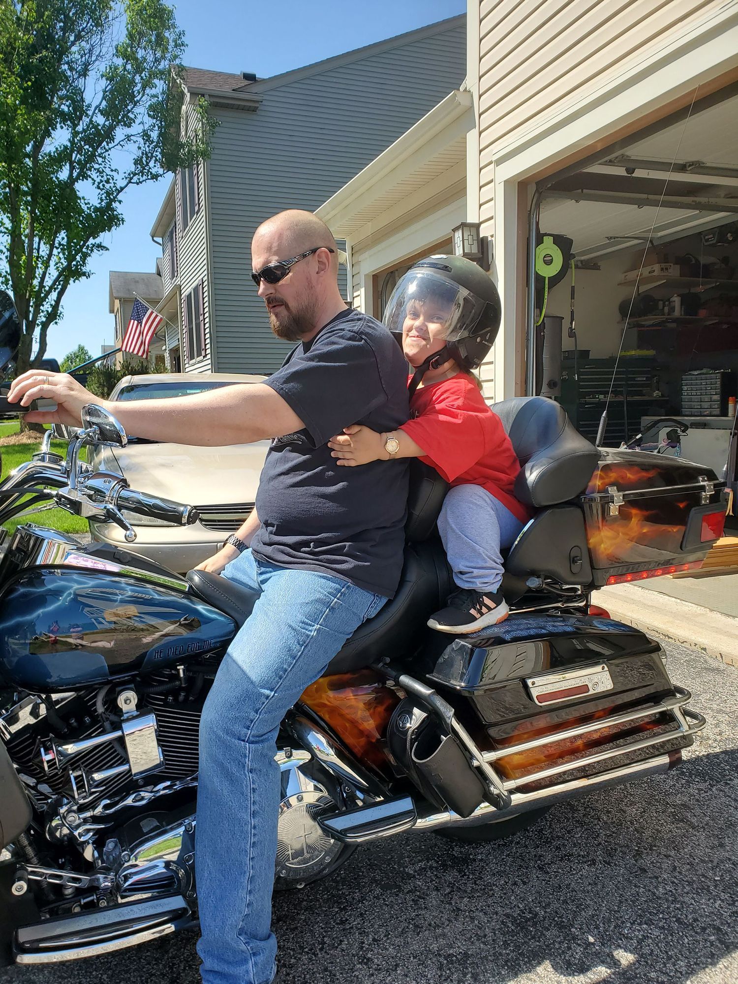 Man on a motorcycle with a child passenger, both wearing helmets, in front of a garage.