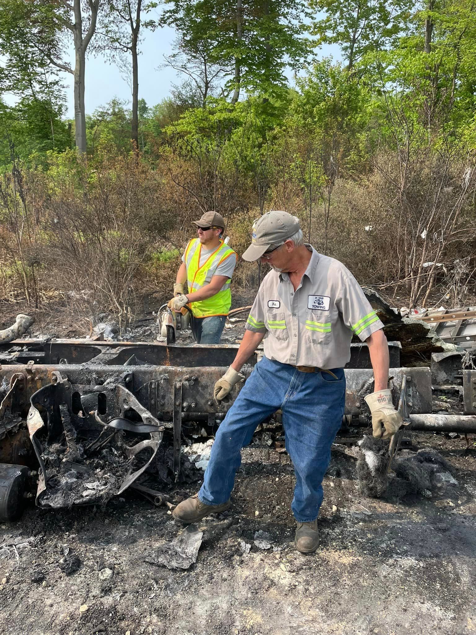 Two workers in safety gear inspecting a burned-out vehicle wreck in a scorched roadside area