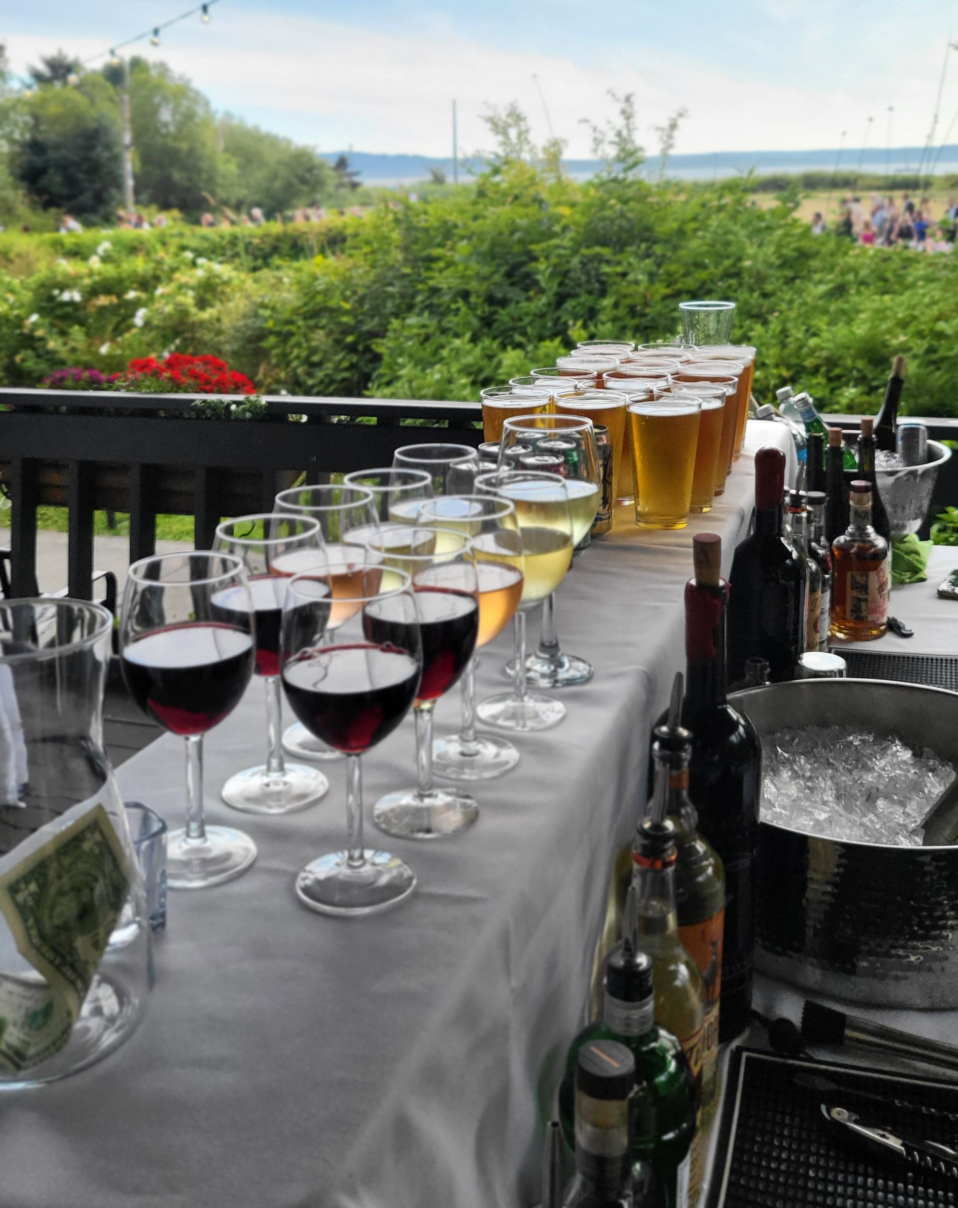 A bar setup outdoors on a deck, featuring rows of wine and beer glasses on a tablecloth with bottles and ice nearby.