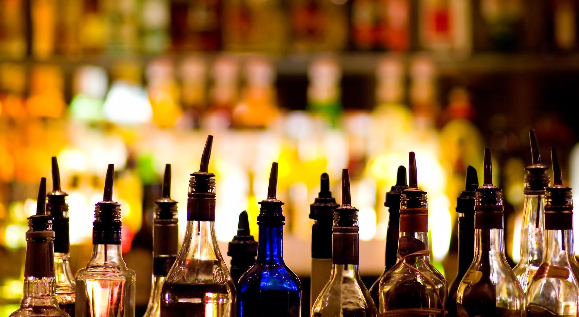 A row of glass liquor bottles with pour spouts against a softly blurred, colorful background of a bar.