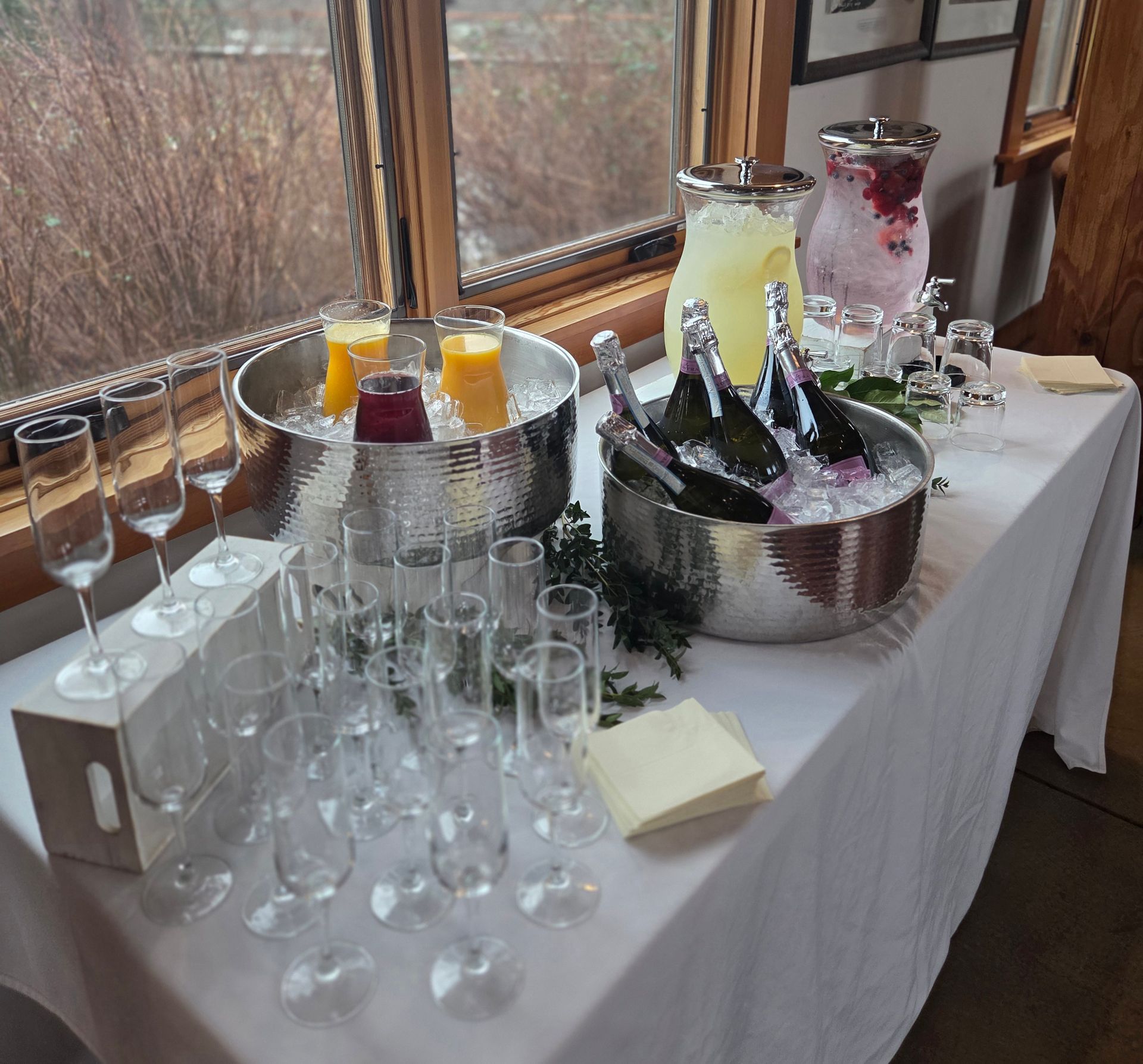 A mimosa bar set up on a white-clothed table with bowls of ice holding champagne bottles, juice, and numerous empty flutes.