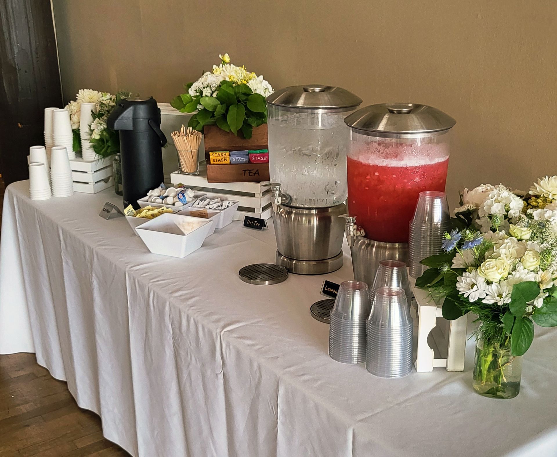 A catering table with coffee, iced water, red punch, and floral arrangements on white tablecloths.