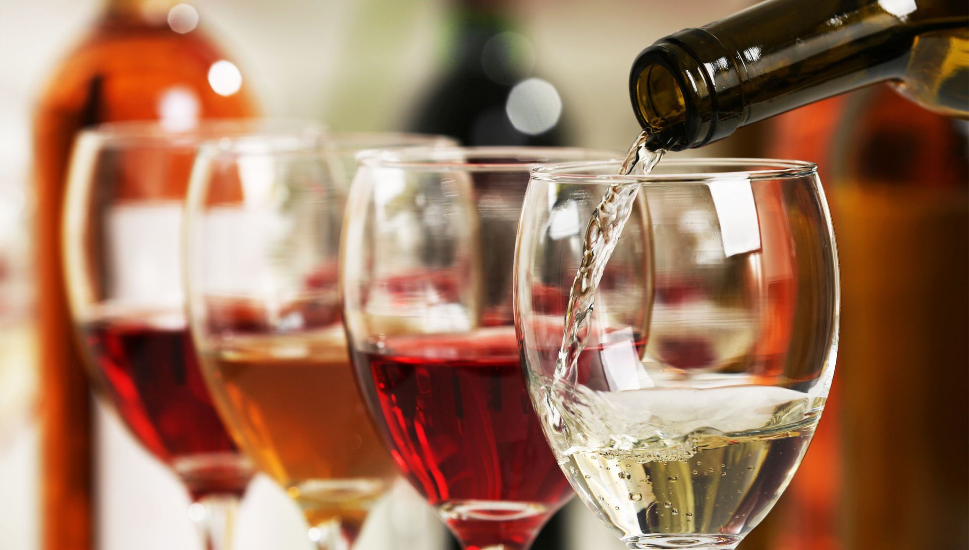 White wine is poured into a glass, with other glasses of white, rosé, and red wine lined up in the background.