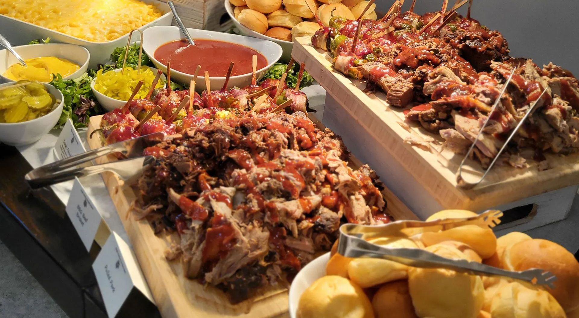 A buffet table featuring platters of BBQ pork, kebabs, sides of pickles, sauce, mac and cheese, and bread rolls.