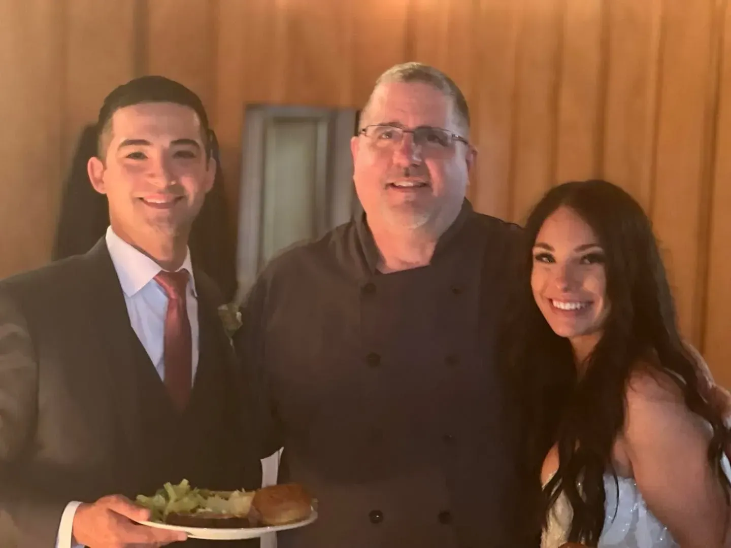 A person in a suit, a chef in a jacket, and a woman smile together in a room with wood paneling.