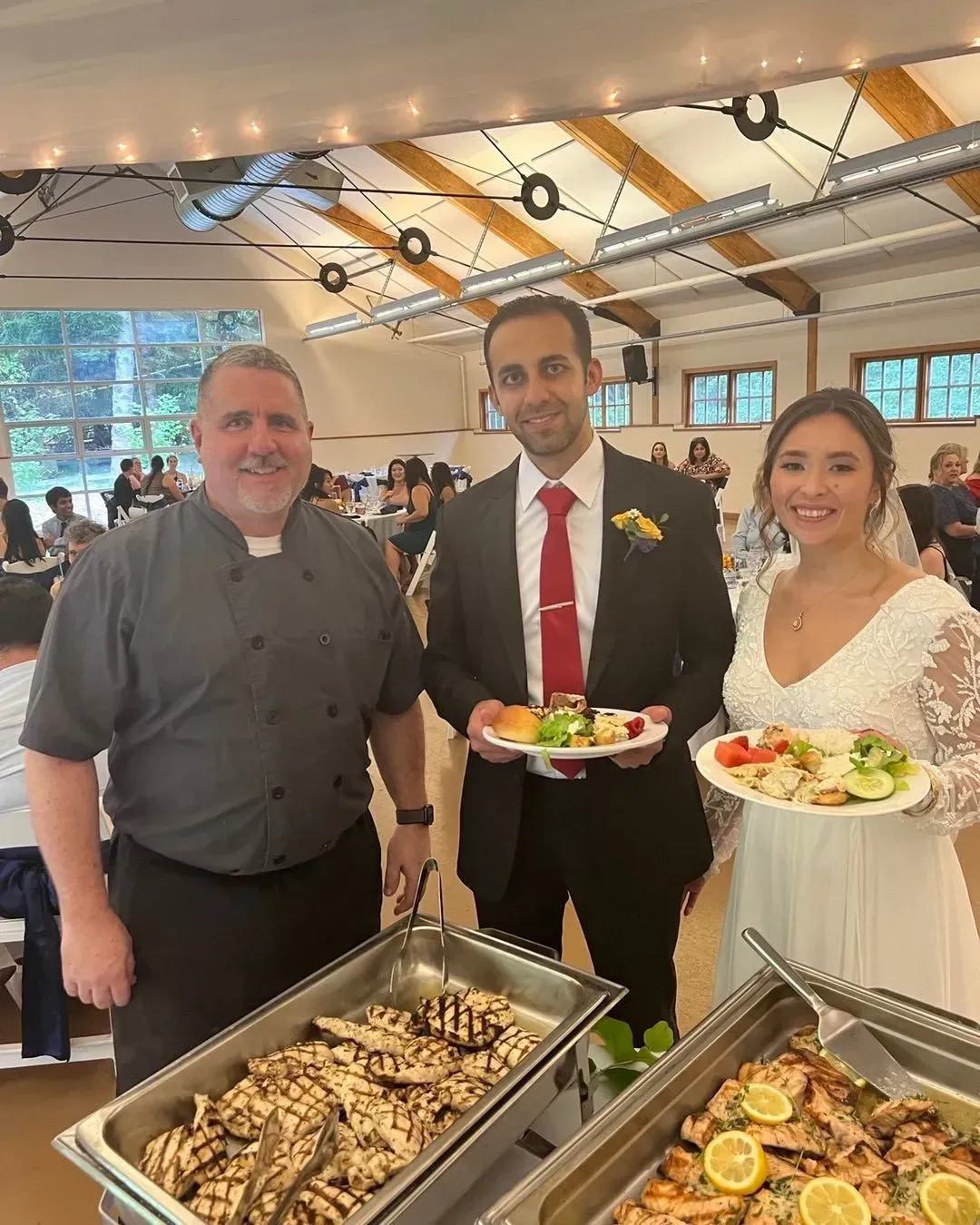 A caterer stands with a smiling bride and groom holding plates of food at a buffet in a bright, rustic event space.