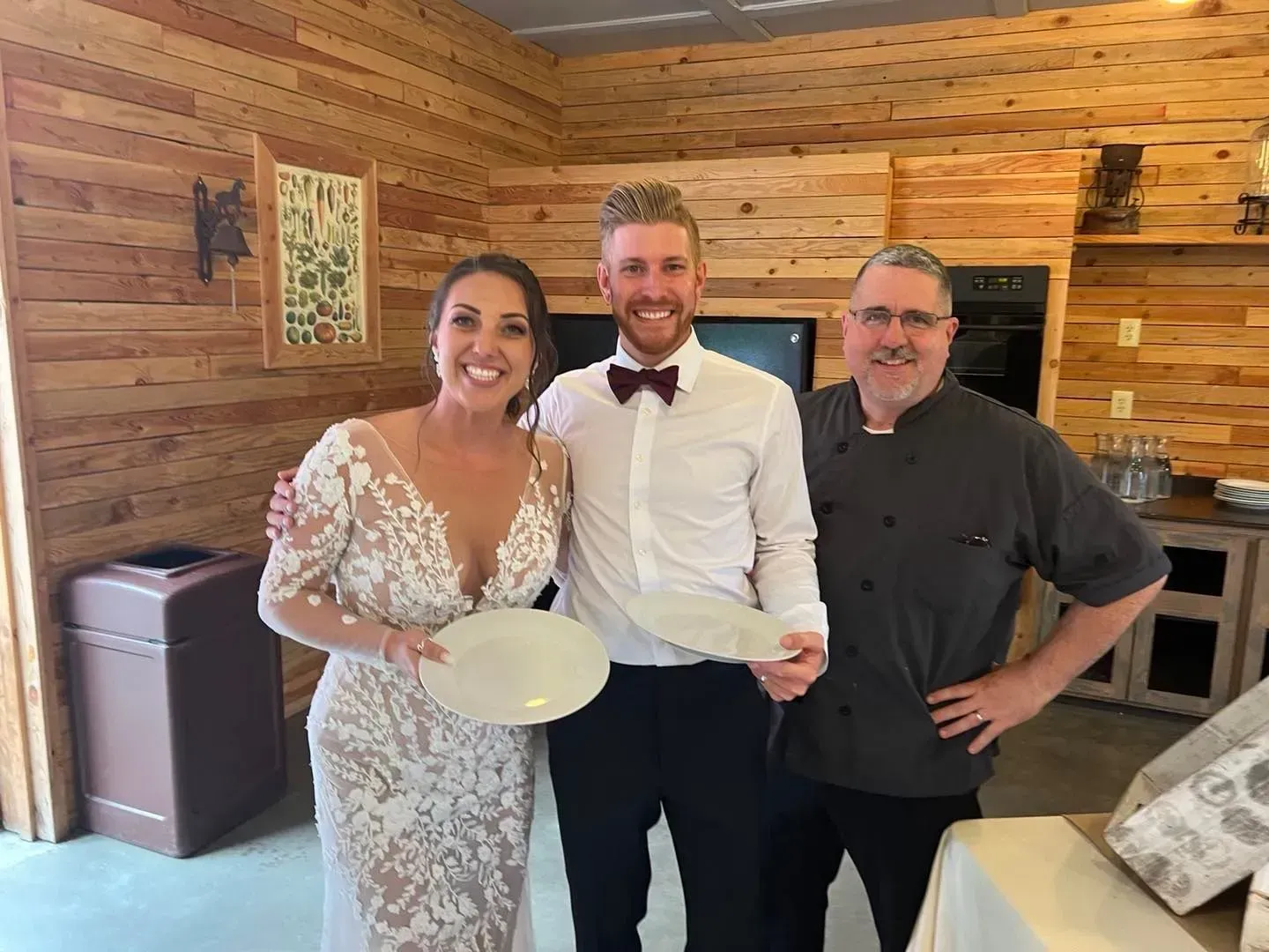 A smiling bride, a man in a formal white shirt with a bowtie, and a chef in a grey uniform hold plates in a wood-paneled room.