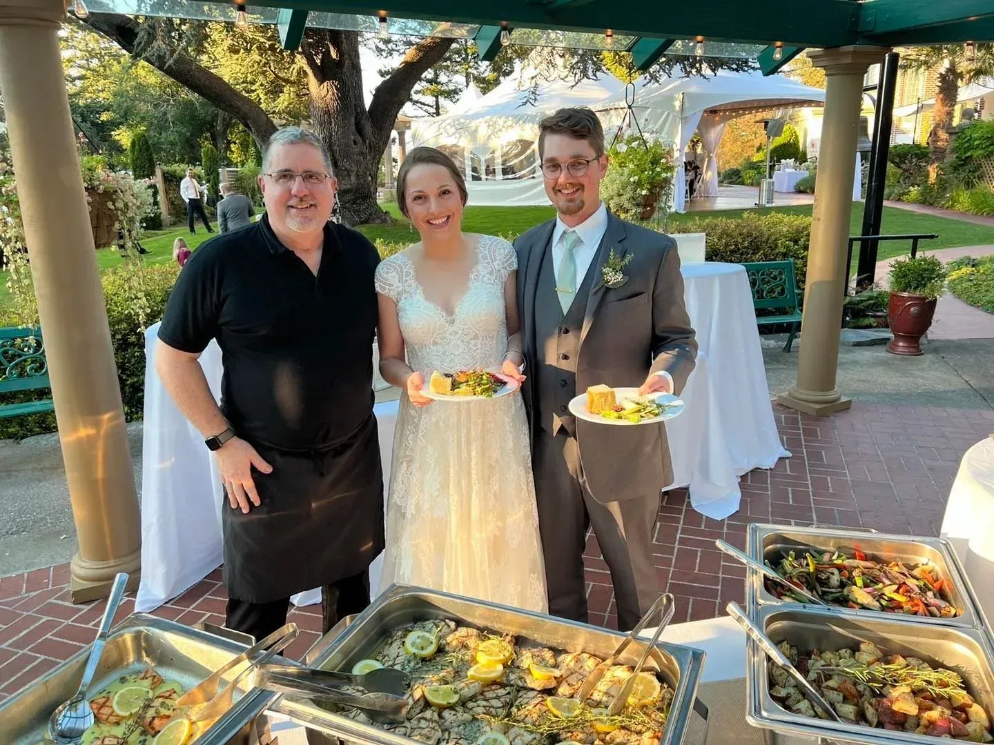 A caterer, a bride, and a groom stand smiling outdoors behind food serving chafing dishes at a wedding reception.