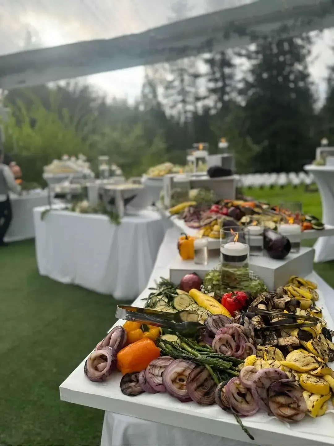 A table spread of grilled, colorful vegetables outdoors on a lawn at a catered event with more tables in the background.
