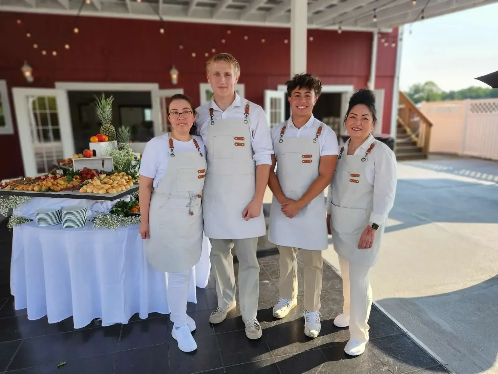 Four catering staff wearing white aprons and shirts smile in front of a food buffet table outside a red building.