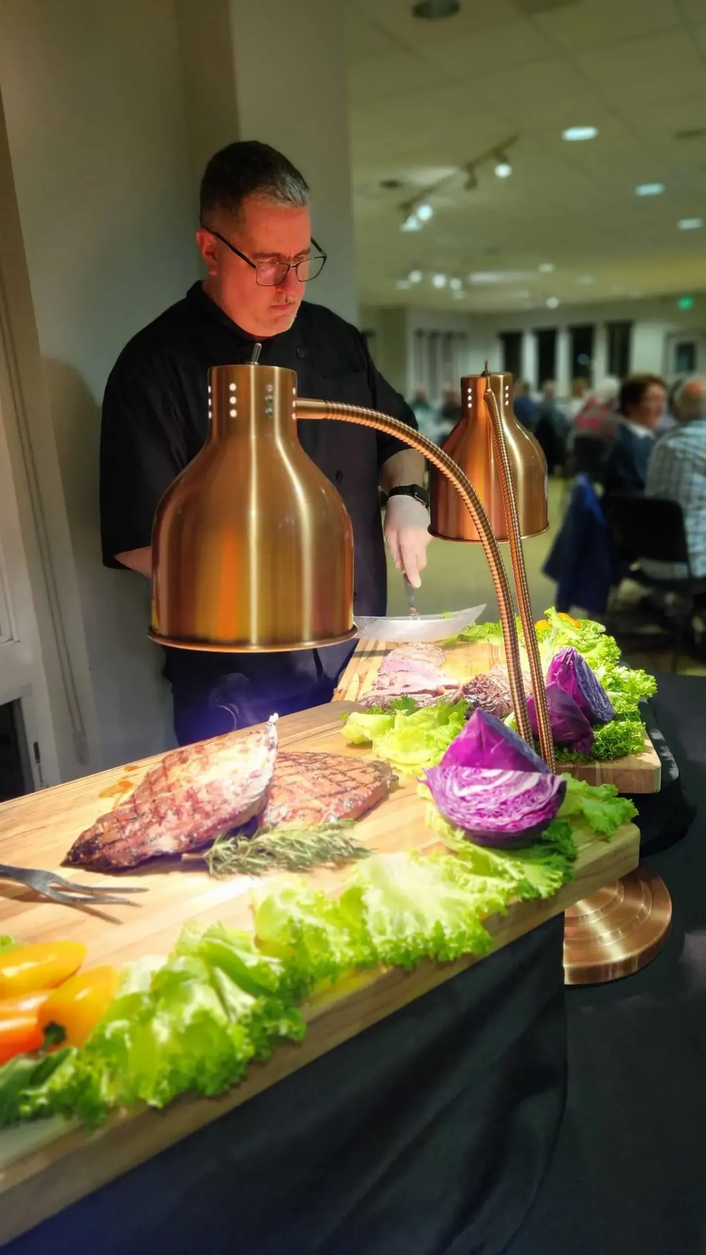A chef carves roasted meat at a buffet station lit by heat lamps, with lettuce and purple cabbage garnish on the table.