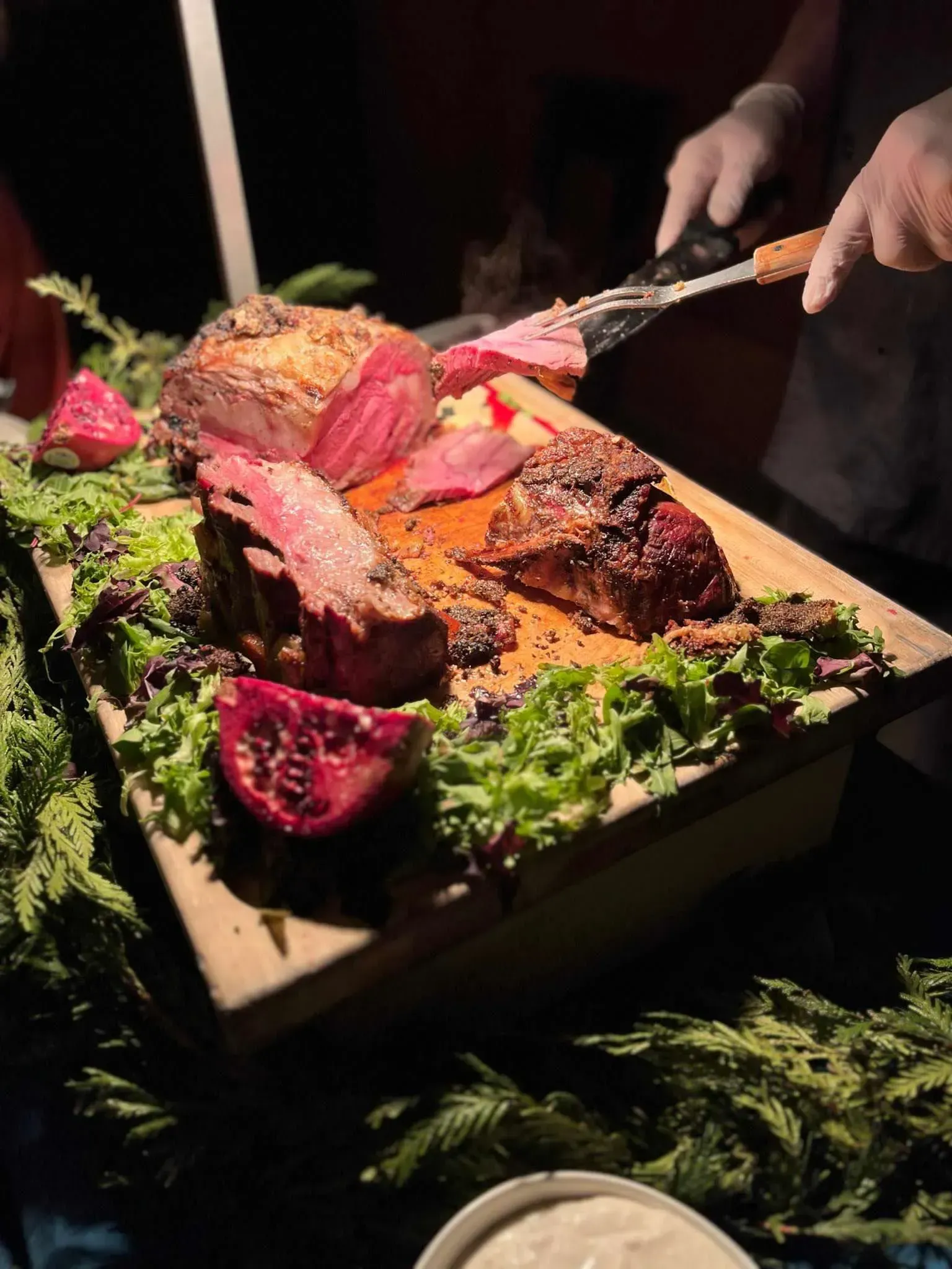 A person wearing gloves carves a thick slice of medium-rare roast beef on a wooden board decorated with greens and fruit.