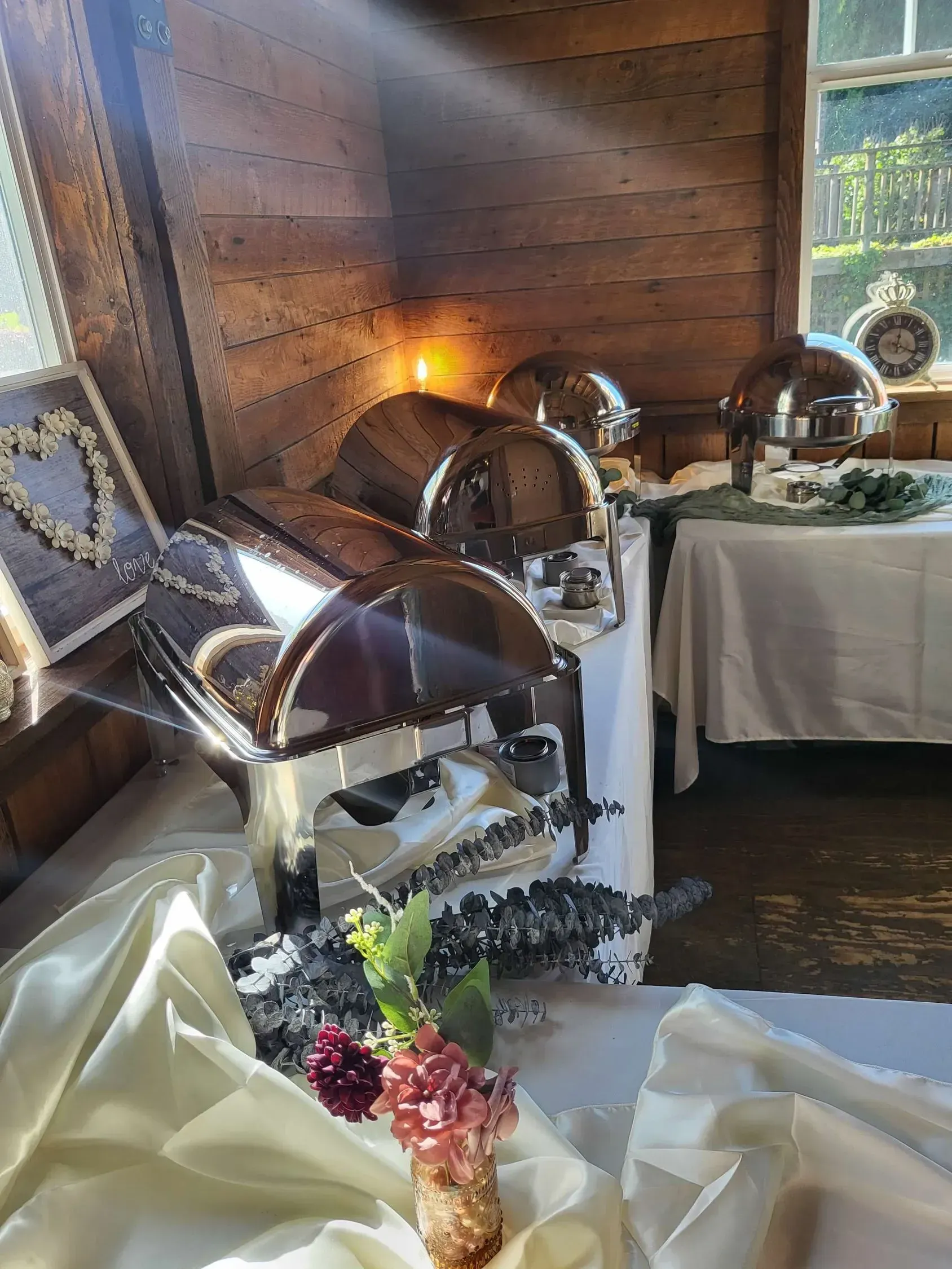 A catering buffet setup in a rustic wooden room with silver chafing dishes on tables covered by white cloths.