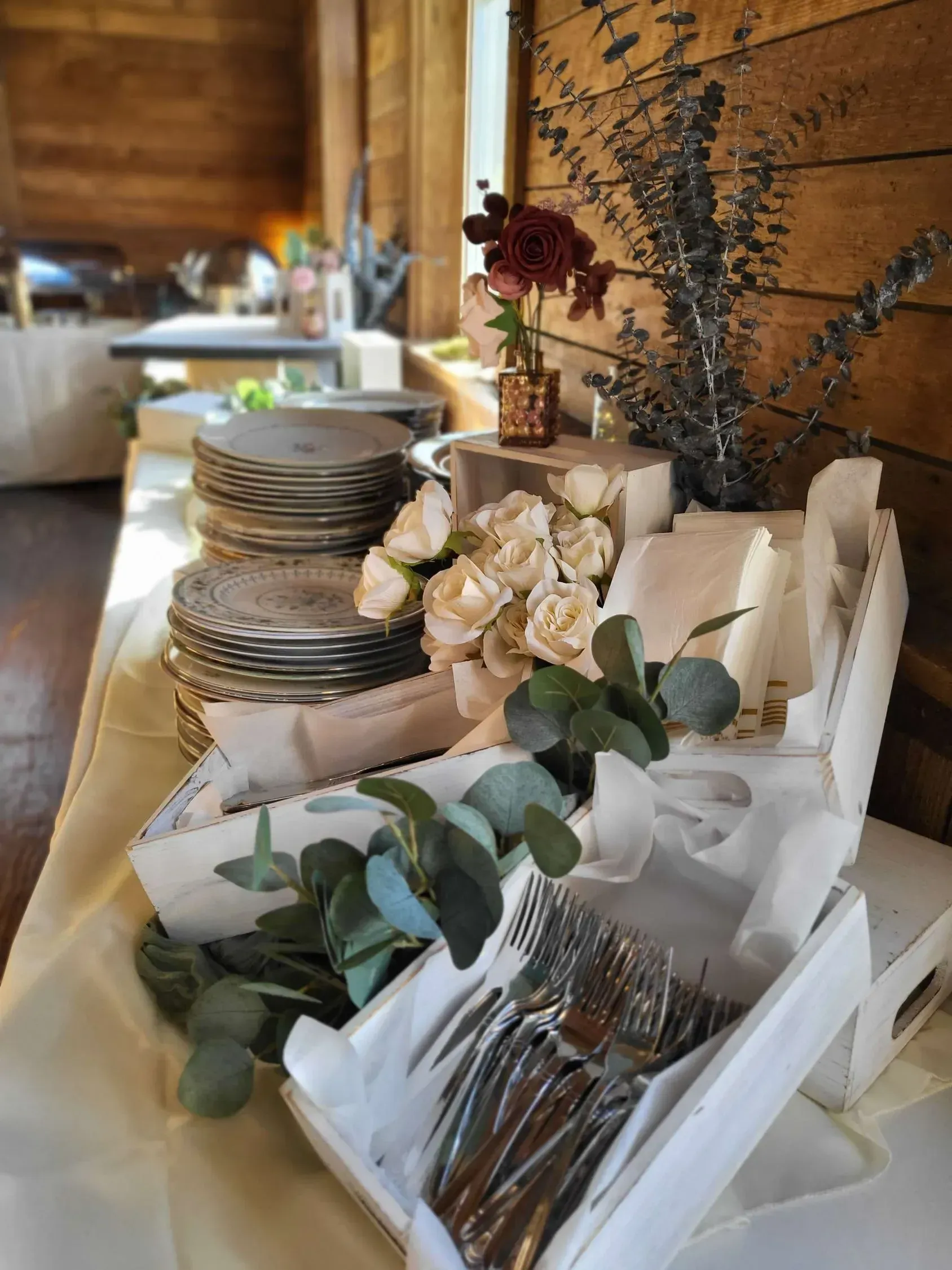 A buffet table set with stacked plates, silverware in white boxes, and floral arrangements against a rustic wood wall.