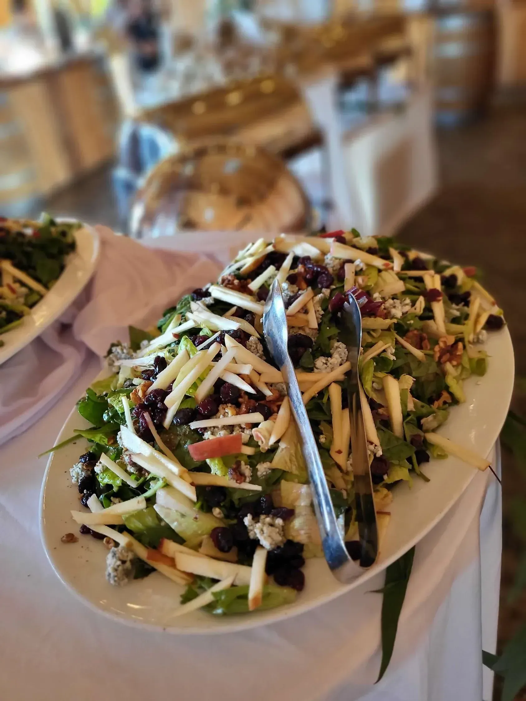 A large oval platter of mixed green salad with sliced apples, dried cranberries, and blue cheese, with tongs included.