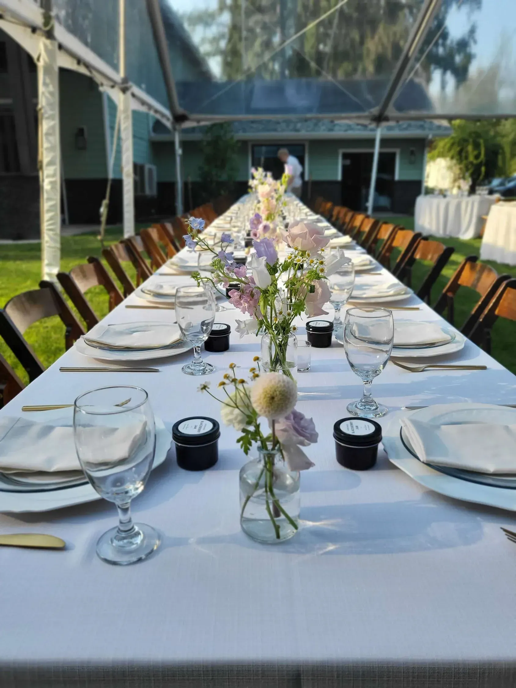 A long banquet table set for a formal outdoor event, featuring white linens, place settings, and floral centerpieces.