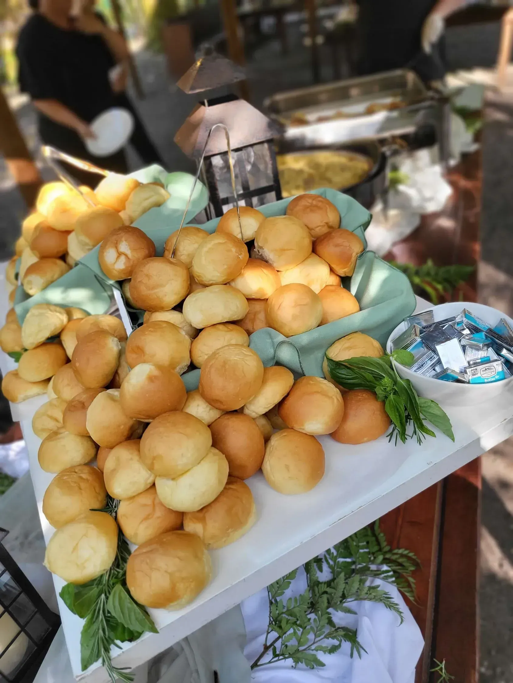 A mound of golden-brown dinner rolls arranged on a white serving tray decorated with green leaves at an outdoor event.