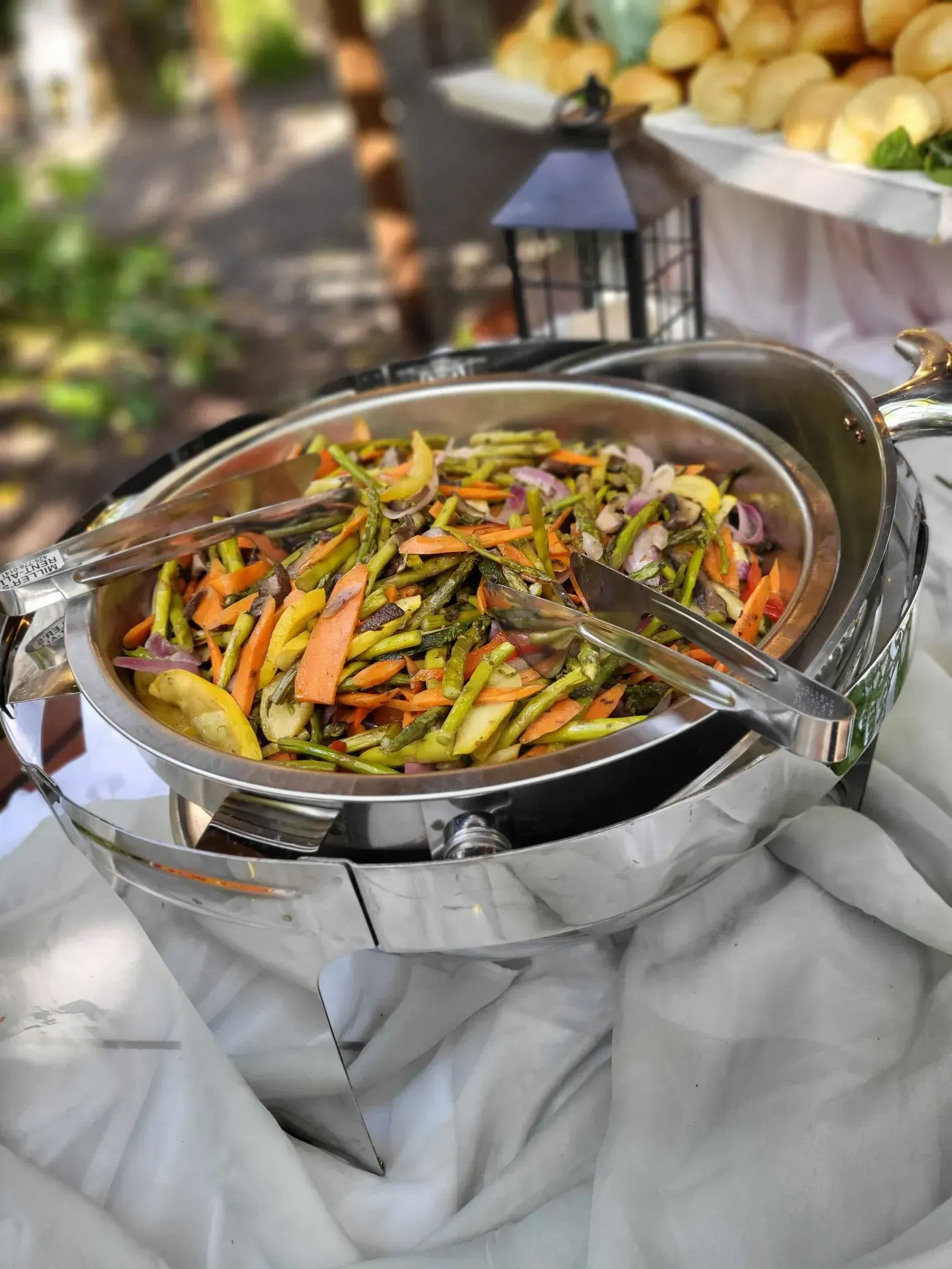A metal chafing dish filled with cooked green beans, sliced carrots, and onions, sitting on a white tablecloth.