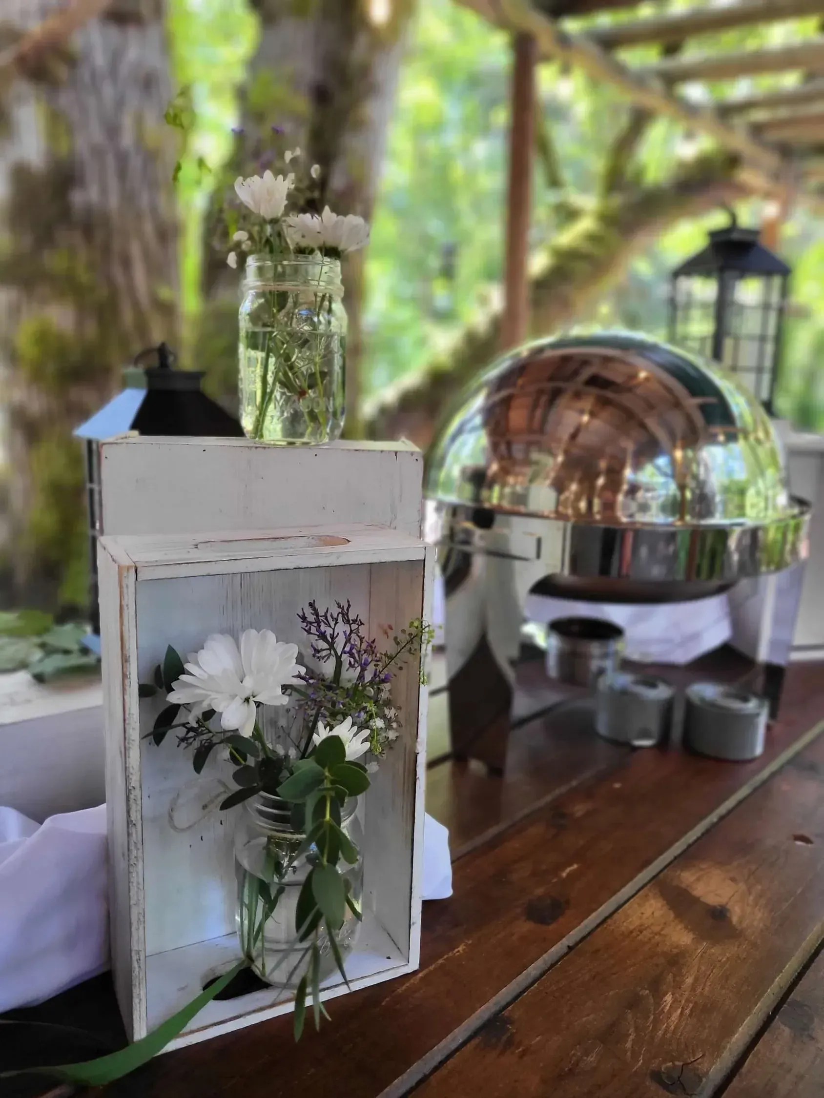 A wooden table decorated with white crates, mason jar flower arrangements, and a silver buffet warmer in a forest setting.