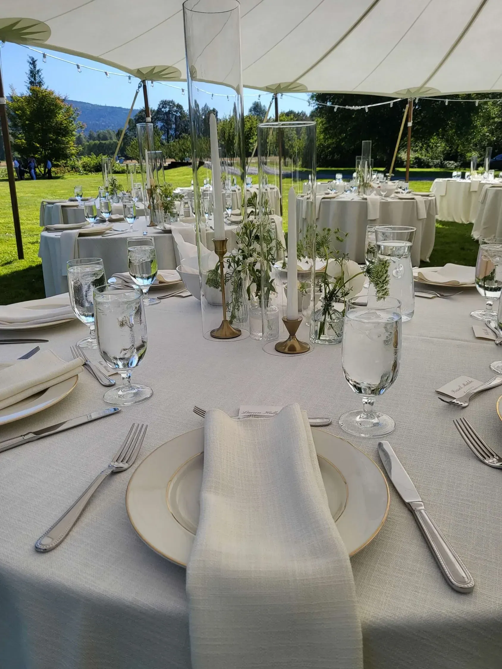 A formal outdoor event setting with round tables draped in cream linens, featuring elegant place settings and floral decor.