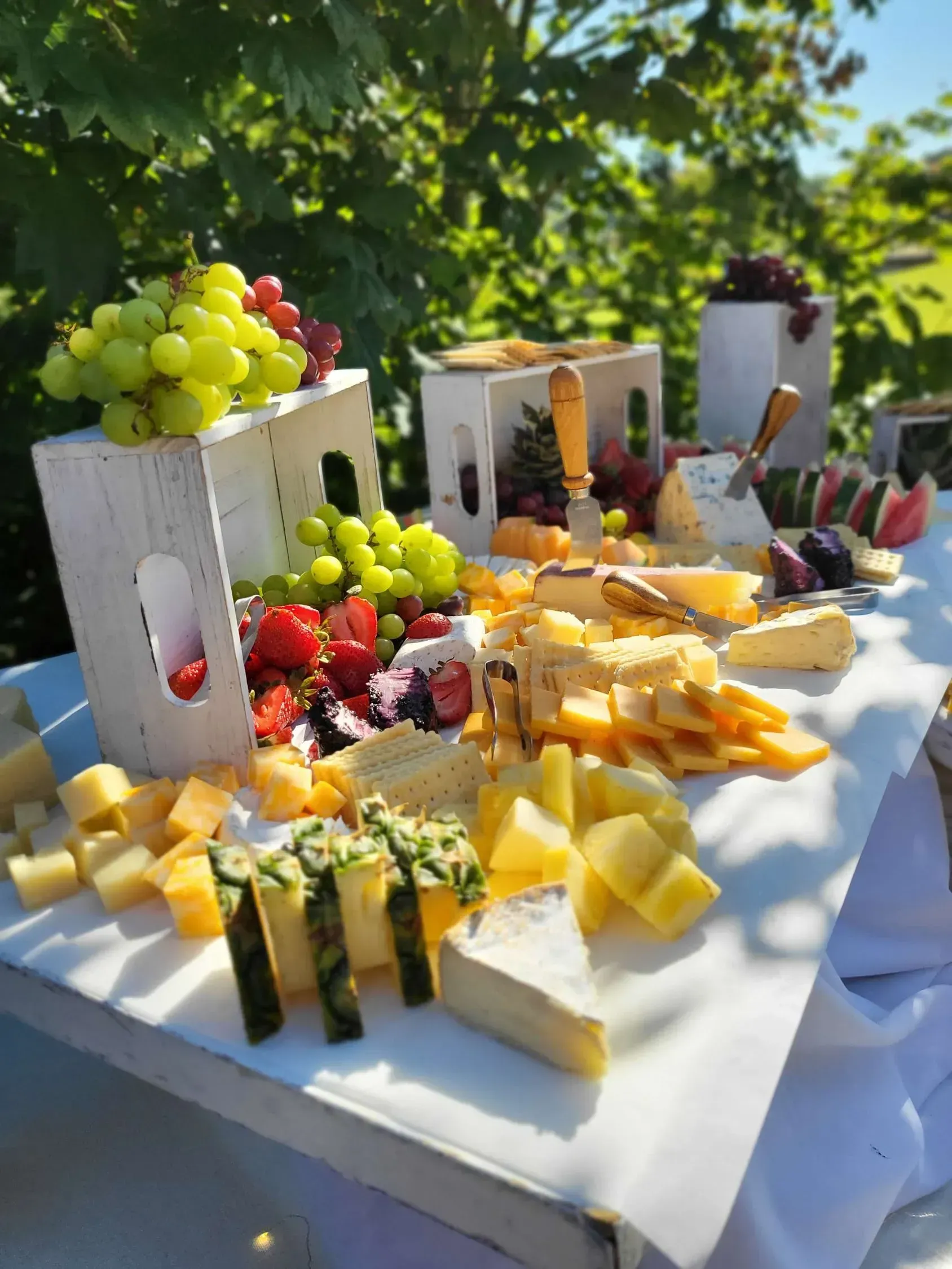 A bountiful charcuterie spread with various cheeses, grapes, and fruit arranged on white wooden crates outdoors.