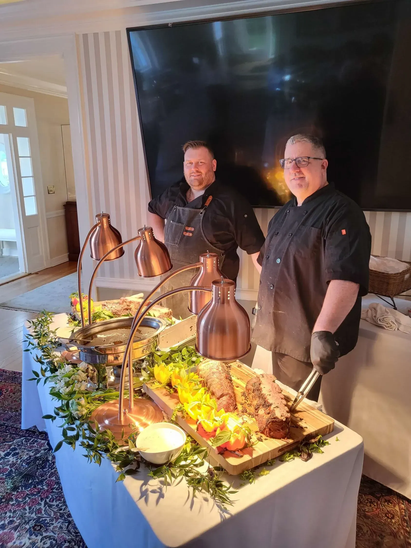 Two chefs in black uniforms prepare a buffet station with warm lighting, foliage, and carved meat on a white tablecloth.
