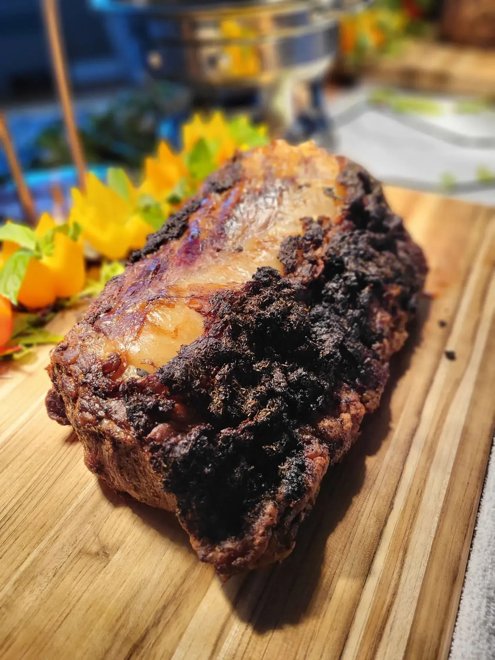 A roasted, charred piece of meat served on a wooden cutting board, garnished with yellow physalis berries.
