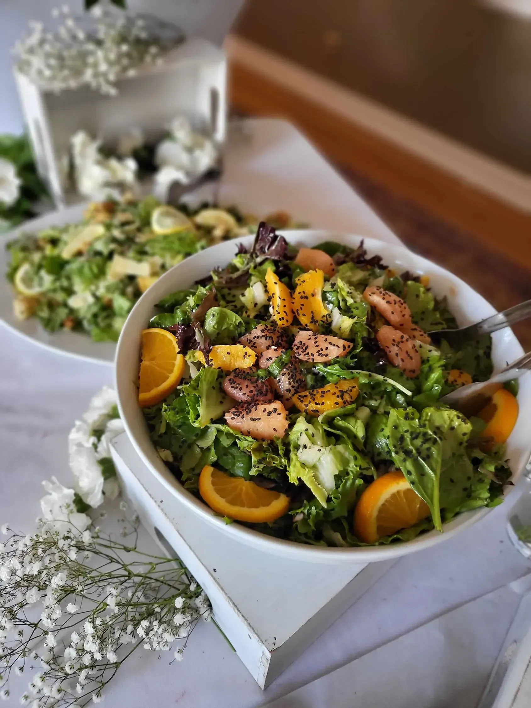 Two bowls of fresh green salad topped with orange slices and seeds, displayed on a table decorated with white flowers.
