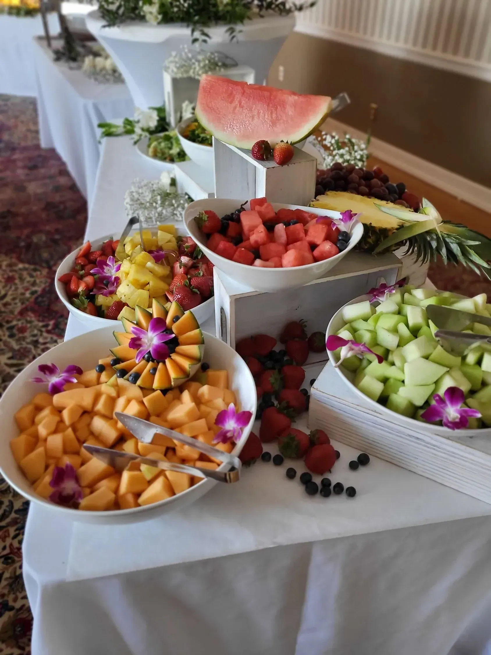 A fruit display on a white tablecloth featuring bowls of cubed melon, watermelon, and pineapple garnished with orchids.