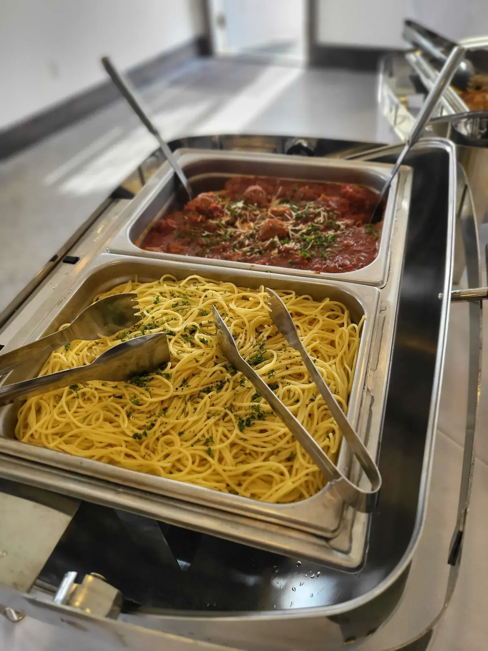 A stainless steel buffet tray with a large portion of spaghetti next to a tray of meatballs in marinara sauce.
