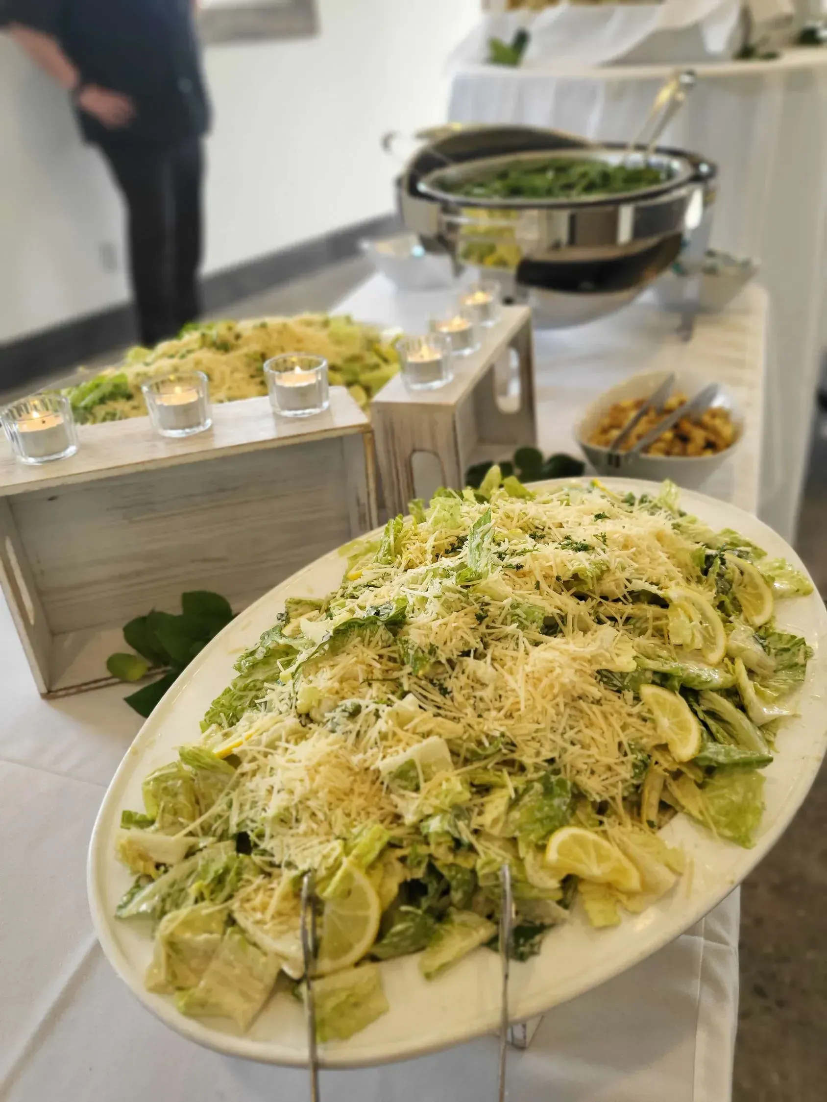 A buffet table features a large oval platter of Caesar salad topped with cheese, beside candles and a chafing dish.