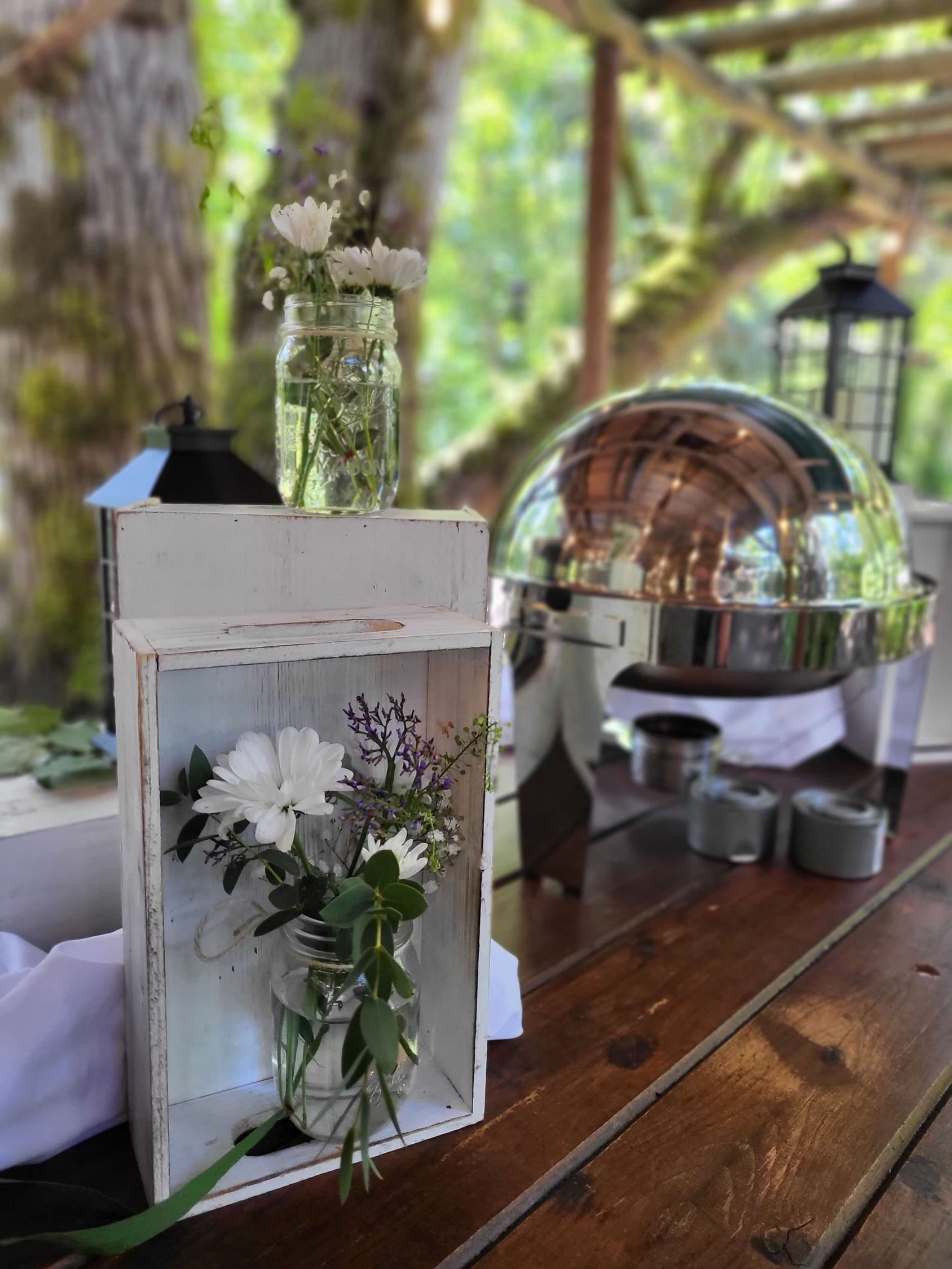White flowers in jars, on a rustic wooden table with a silver buffet and lanterns in an outdoor setting.