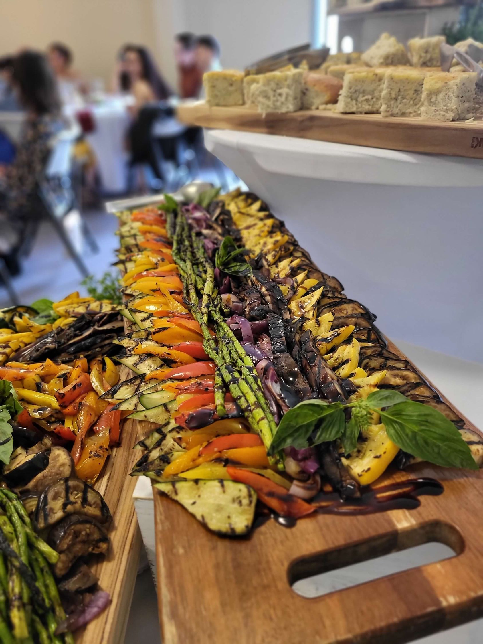 Wooden board with grilled vegetables: peppers, asparagus, zucchini. People in the background.