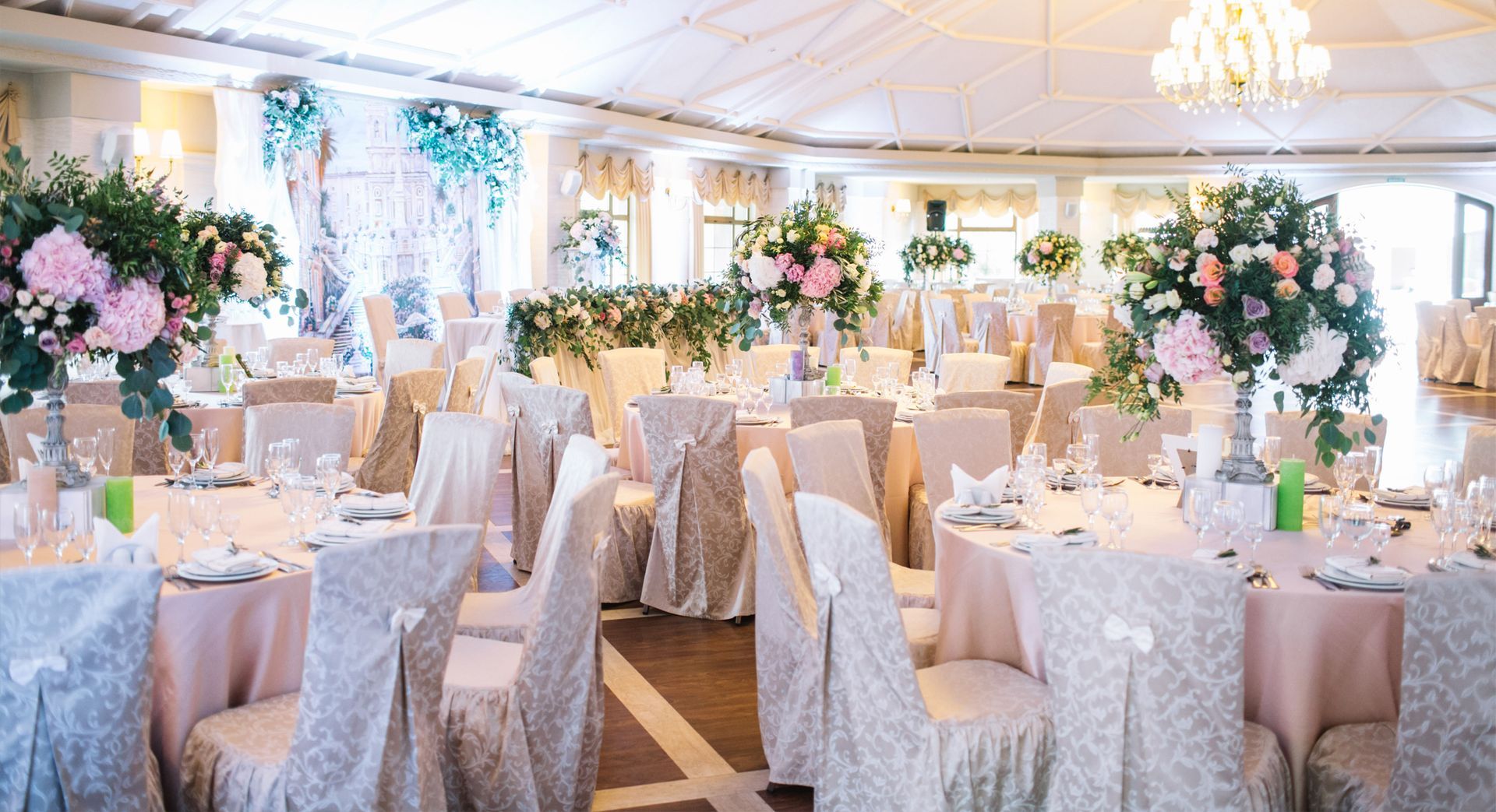 A bright banquet hall decorated for a wedding with round tables, light pink tablecloths, and large floral centerpieces.