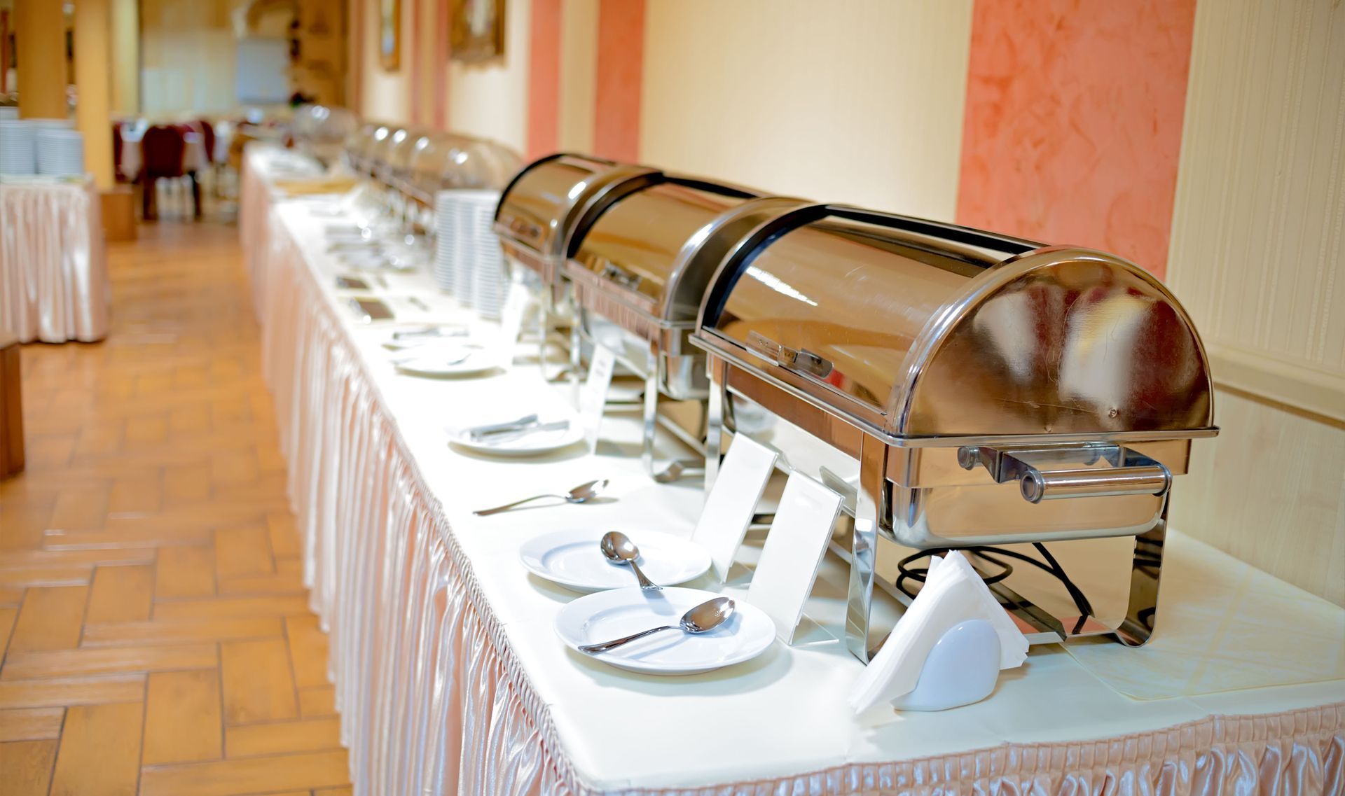 A long buffet table set with shiny metal chafing dishes, white plates, and cutlery in a dining room.