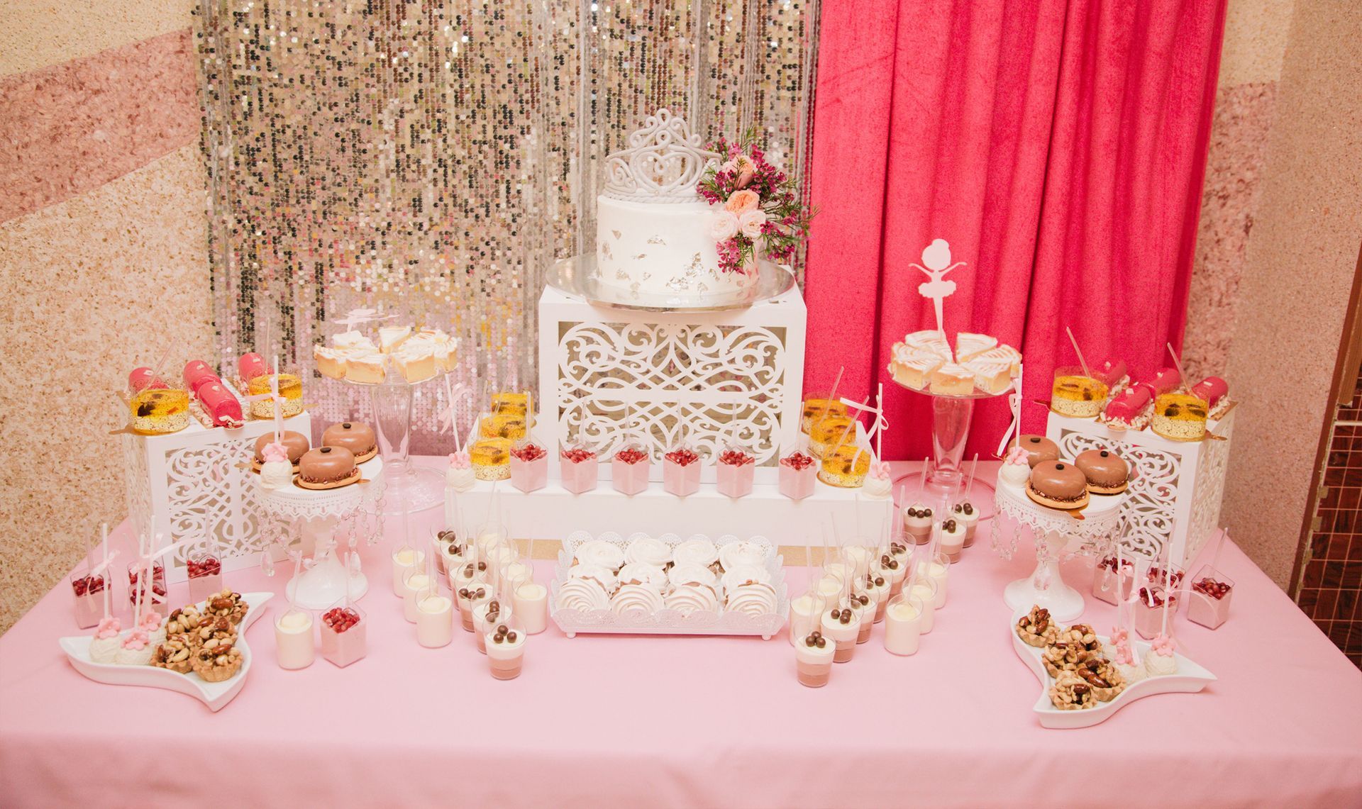 A table covered with a pink tablecloth displaying a tiered cake, various small desserts, and treats in a festive setting.