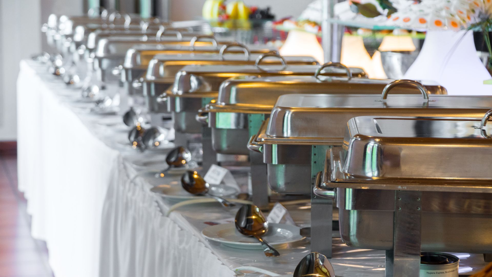 A row of stainless steel catering chafing dishes lined up on a white tablecloth at a buffet.
