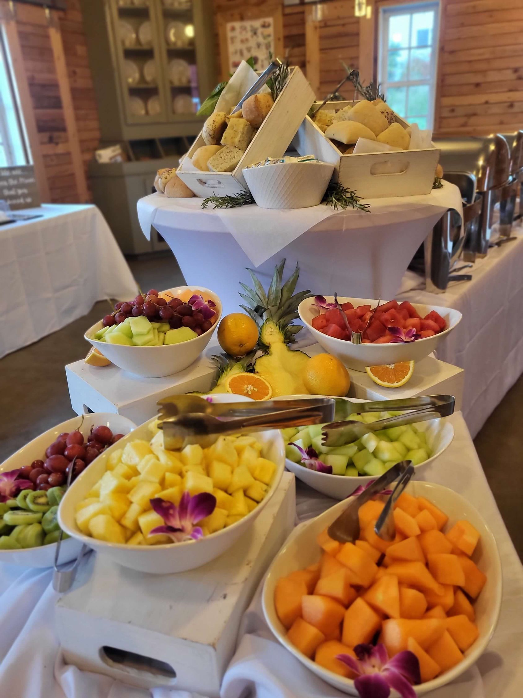 Buffet table with fruit and bread. White bowls hold cantaloupe, pineapple, grapes, and citrus, with serving utensils.
